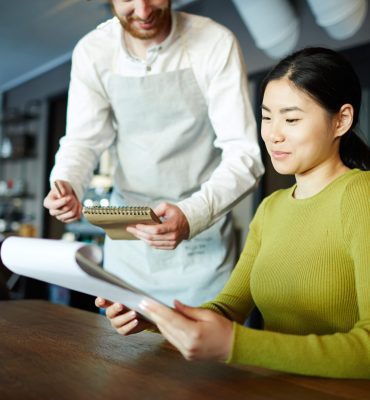 Asian girl visiting restaurant and looking through menu with waiter standing near by